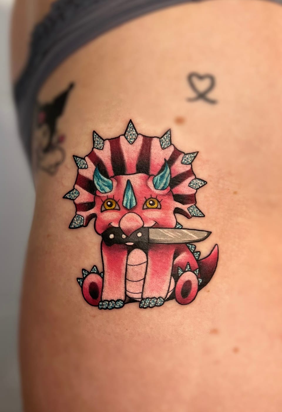 A pink neo-traditional triceratops with teal horns holds a knife in its mouth on an outer thigh.