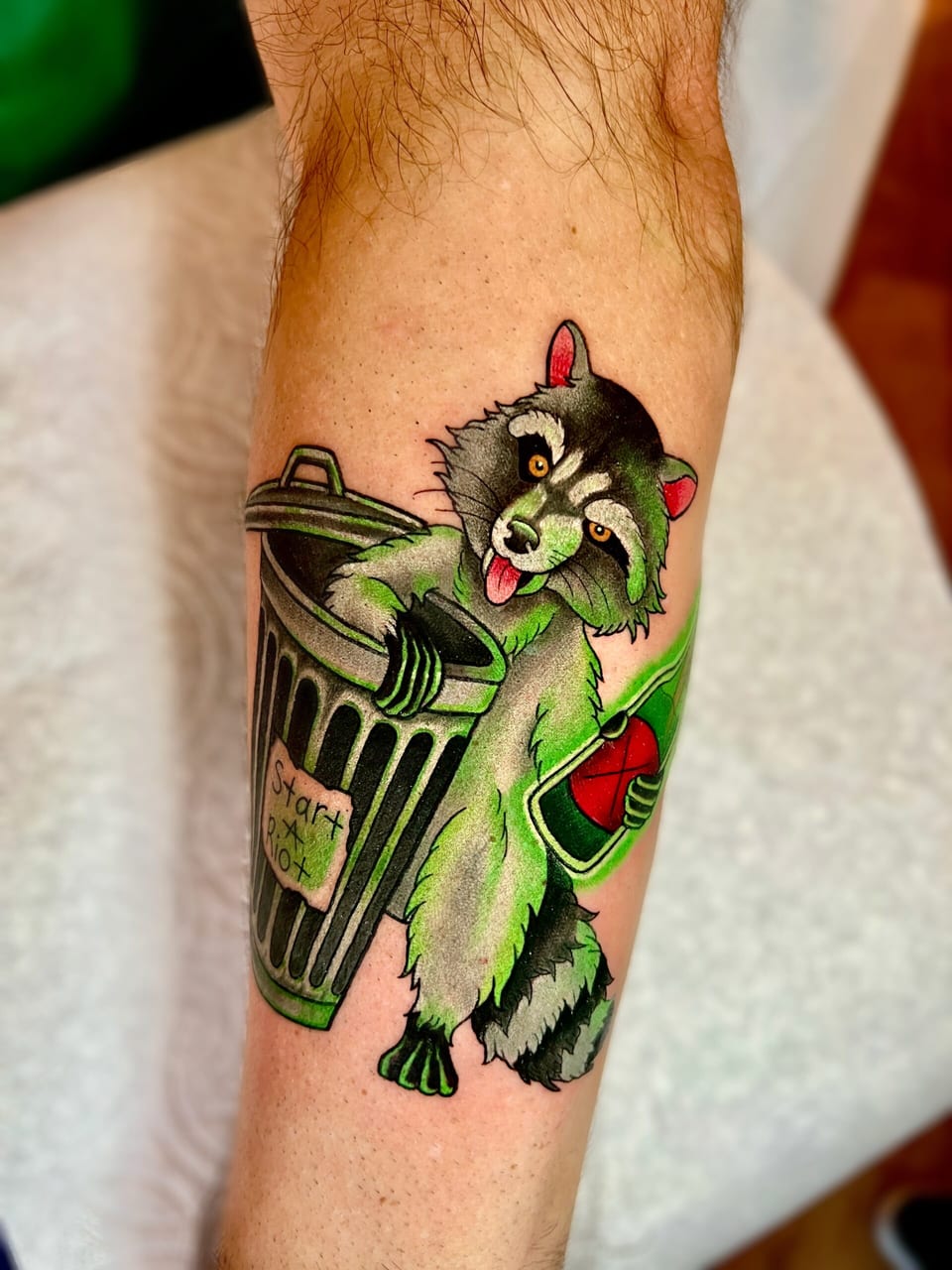 A color neo-traditional raccoon with green shading holds a bottle beside a trash can on a forearm.