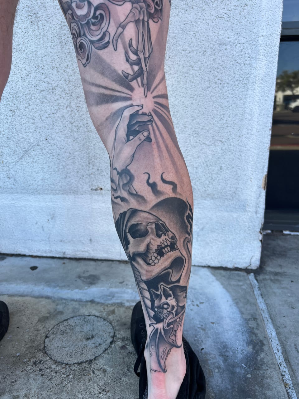 A black and grey illustrative sleeve of reaching hands, rays, a hooded skull and flames runs down a leg.