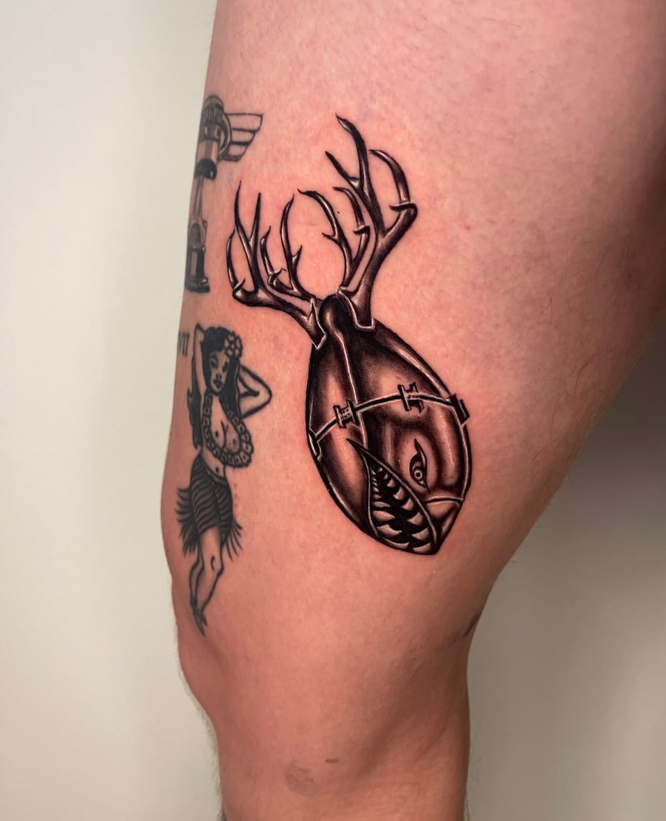 A black and grey illustrative bomb with antlers and a shark grin sits beside a pin-up on an outer thigh.