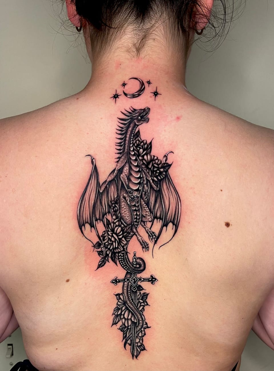 A black and grey illustrative dragon coils around flowers and a sword beneath a crescent moon down the spine.