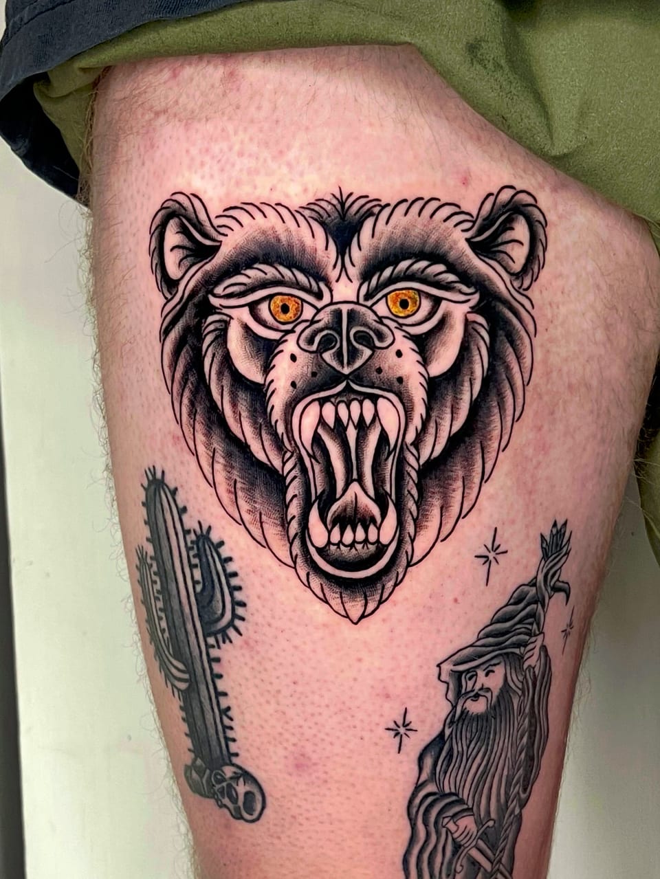 A black and grey neo-traditional bear head with orange eyes and open jaws sits above a cactus and wizard on a thigh.