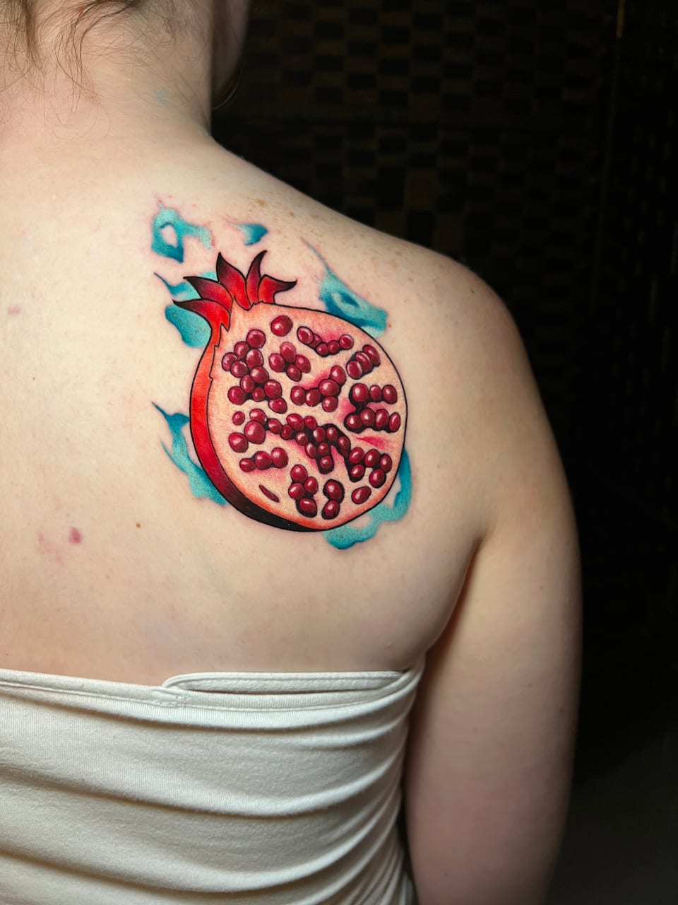 A red neo-traditional pomegranate with exposed seeds and teal flames sits on a shoulder blade.