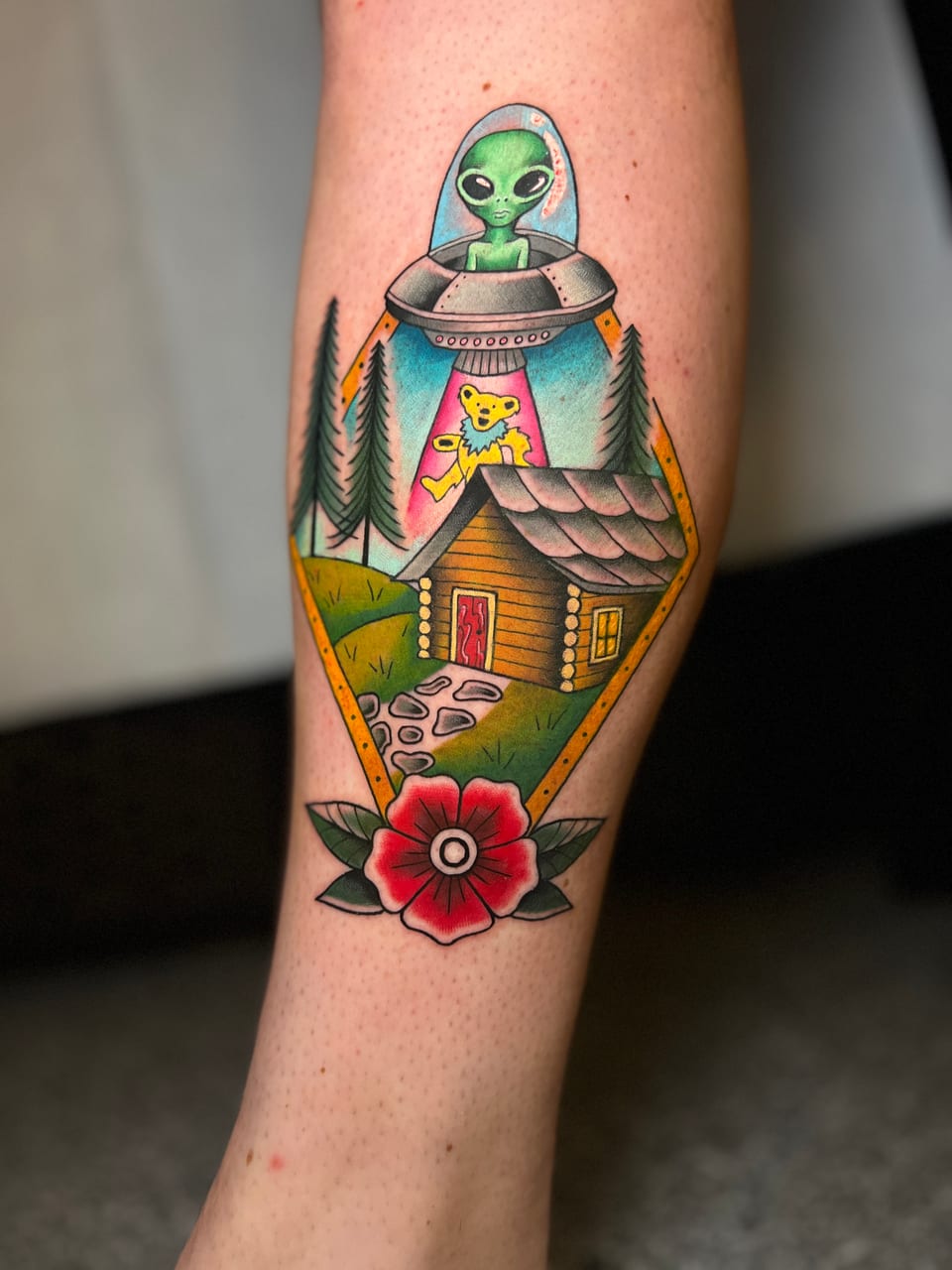 A color traditional alien beams up a bear above a cabin, pine trees and a red flower on an outer forearm.