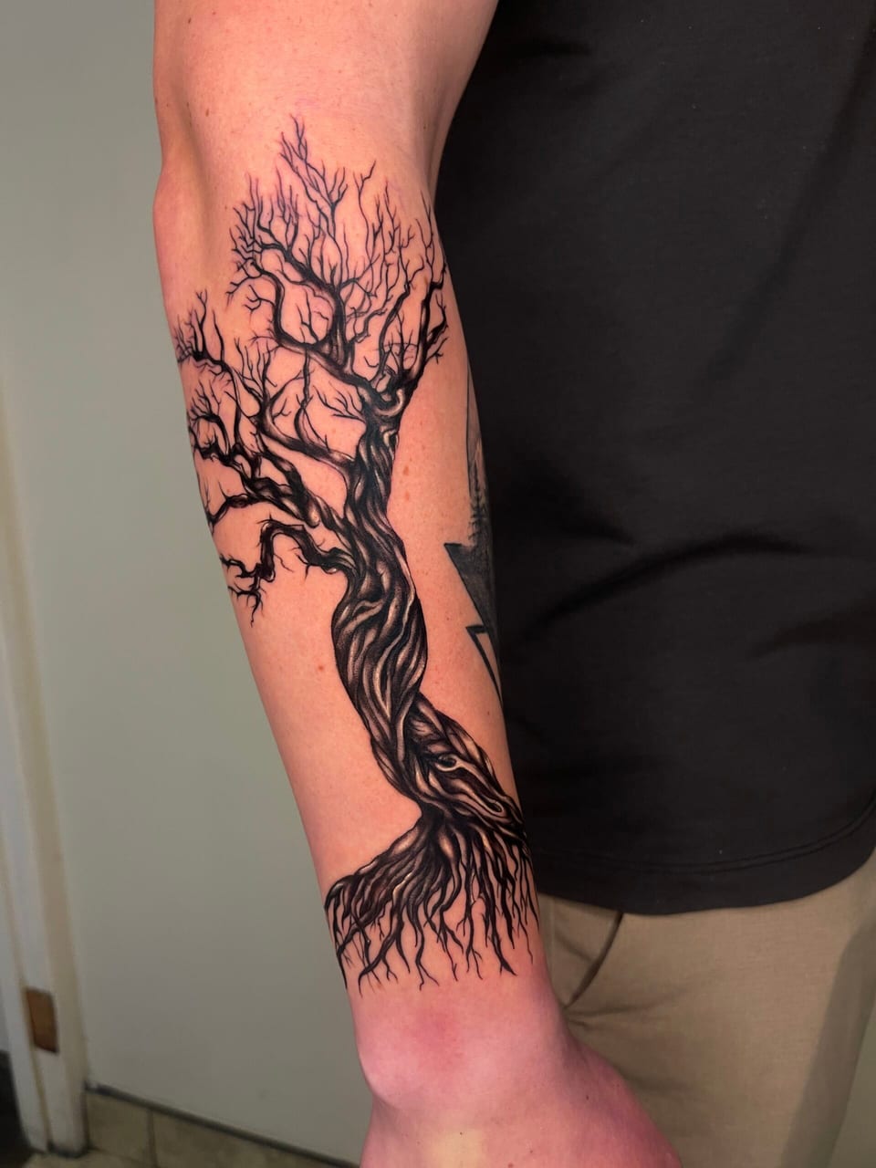 A black and grey illustrative bare tree with a twisted trunk and spreading roots runs down an outer forearm.