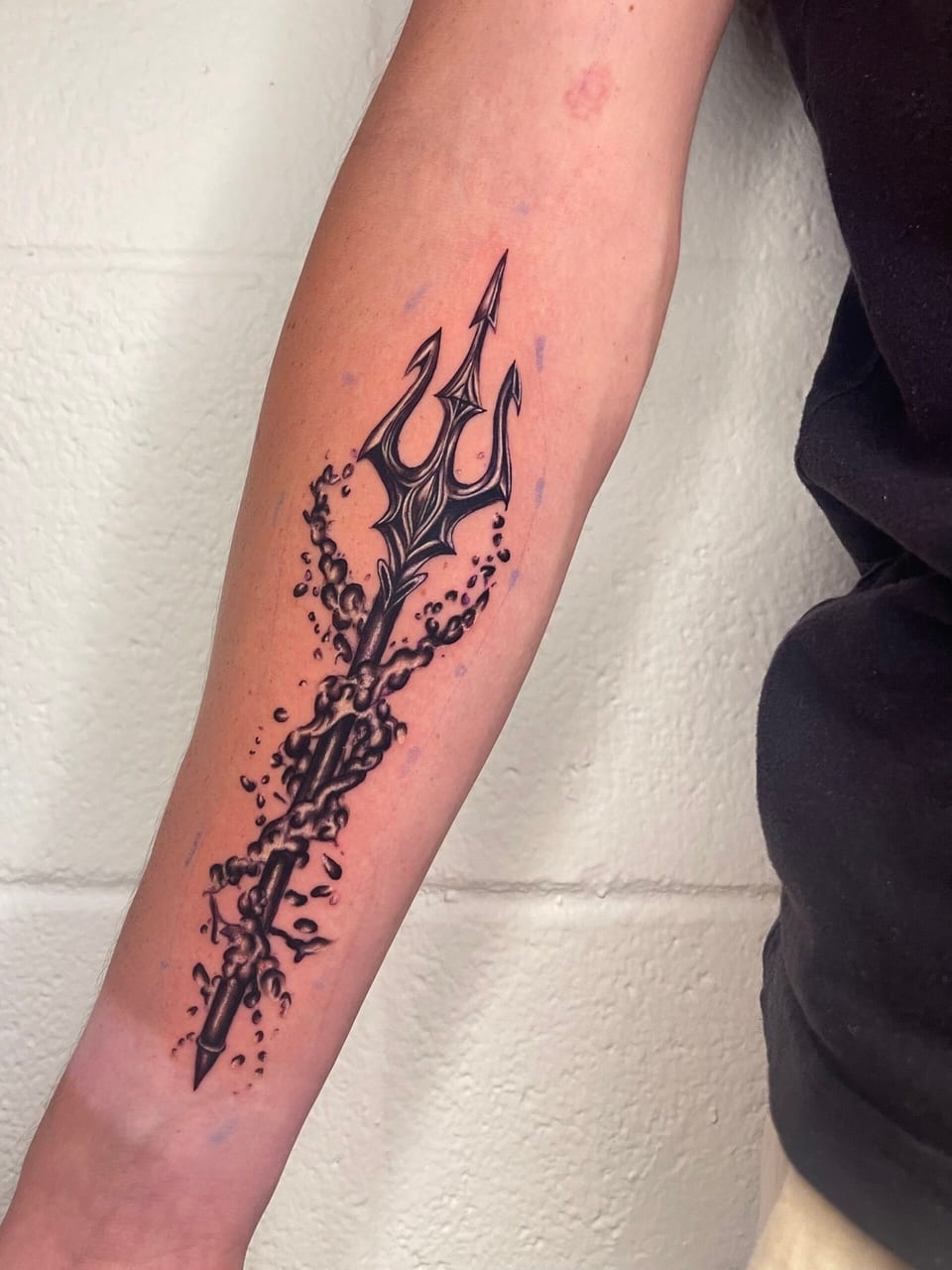 A black and grey illustrative trident surrounded by swirling water and droplets runs down an inner forearm.