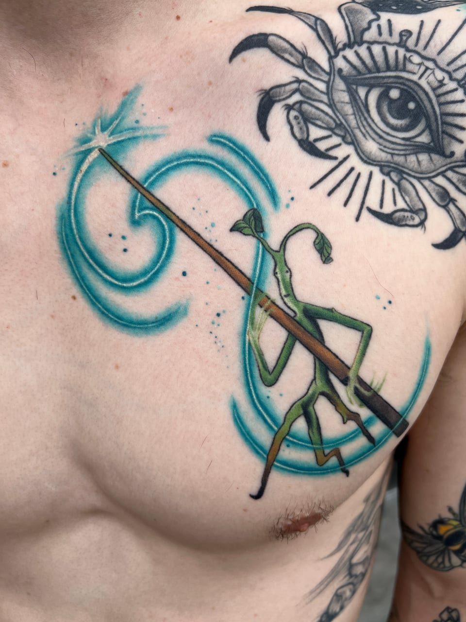 A color illustrative praying mantis holds a staff amid swirling teal watercolor strokes across an upper chest.