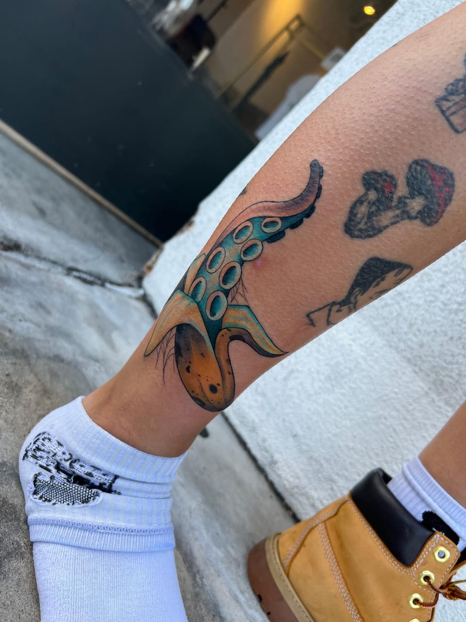A teal neo-traditional octopus tentacle emerges from a yellow banana peel beside mushrooms on an ankle.