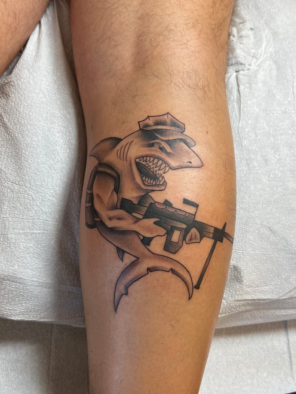A black and grey illustrative shark in a cap holds a rifle on a calf.
