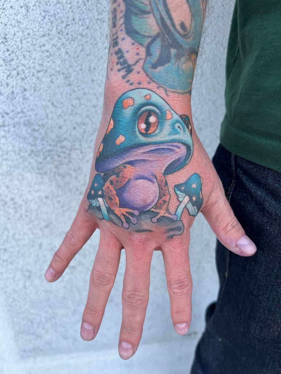 A color neo-traditional frog sits beneath a large teal mushroom among smaller mushrooms on the back of a hand.