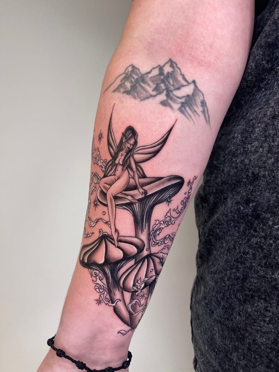 A black and grey illustrative winged fairy sits on mushrooms amid stars and wisps beneath mountains on a forearm.