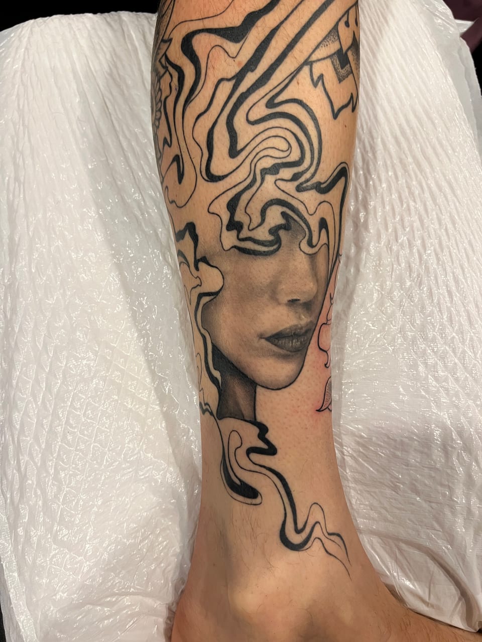 A black and grey realistic woman's face dissolves into flowing blackwork lines down a lower leg.