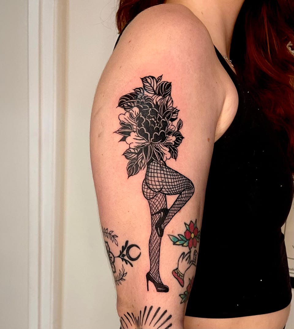 A blackwork pin-up figure with flowers for a head and fishnet stockings stands on an outer upper arm.
