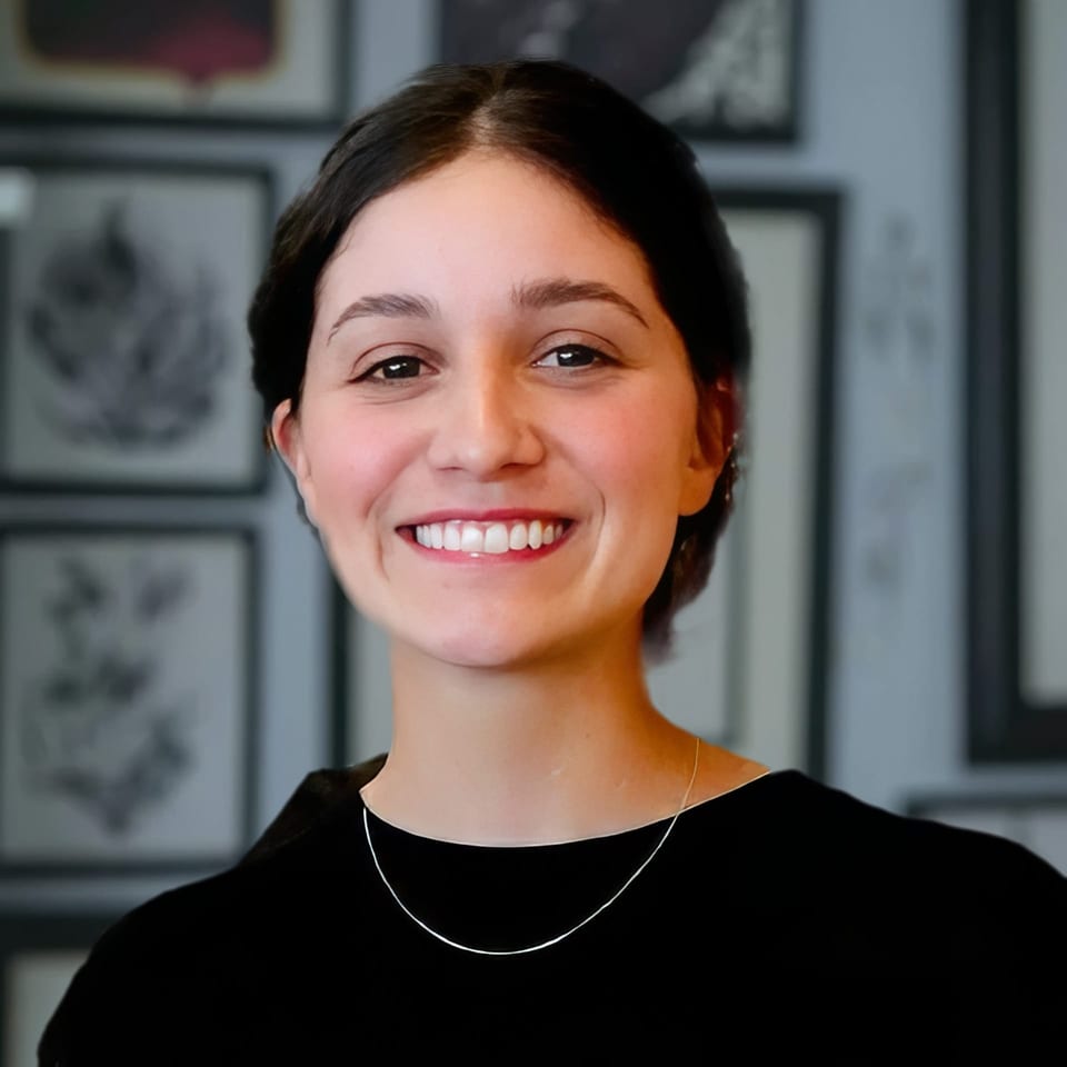 Stellie smiles in a black top and necklace in front of framed artwork.
