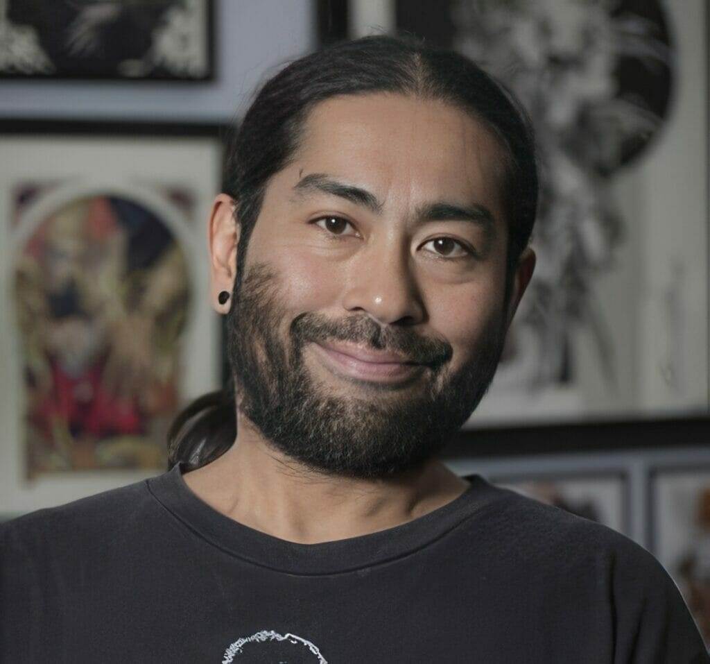 A man with a beard standing in front of framed pictures at a tattoo shop in San Diego.
