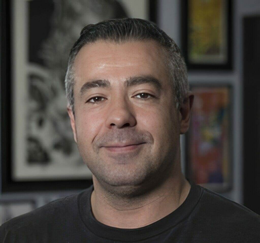 A man in a black shirt smiling in front of framed pictures at a tattoo shop.