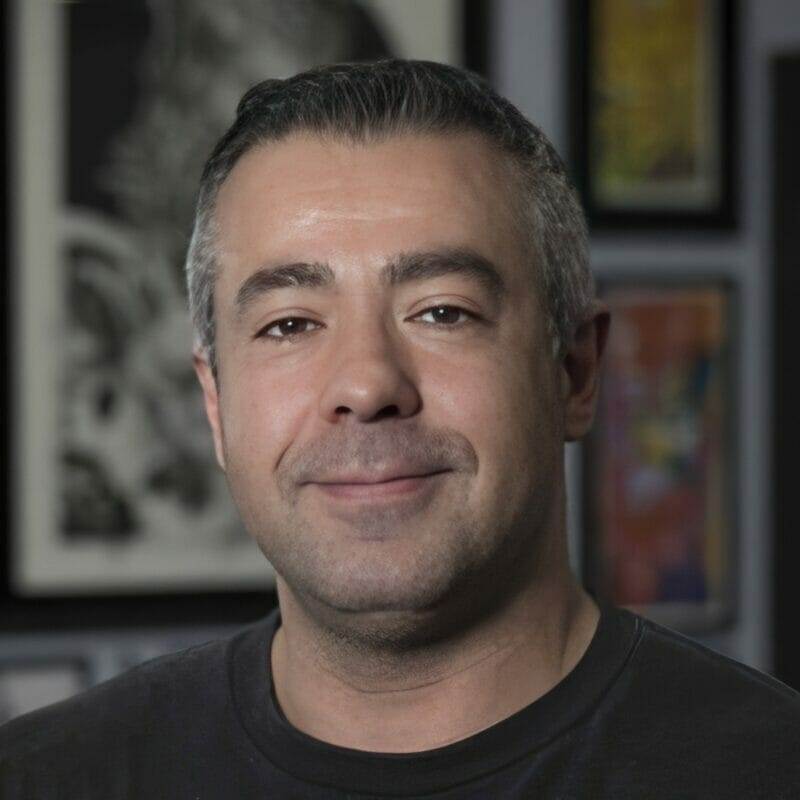 A man in a black shirt smiling in front of framed pictures at a tattoo shop.