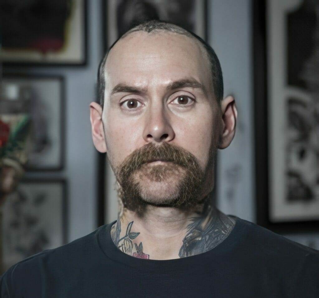 A man with tattoos in front of a wall.