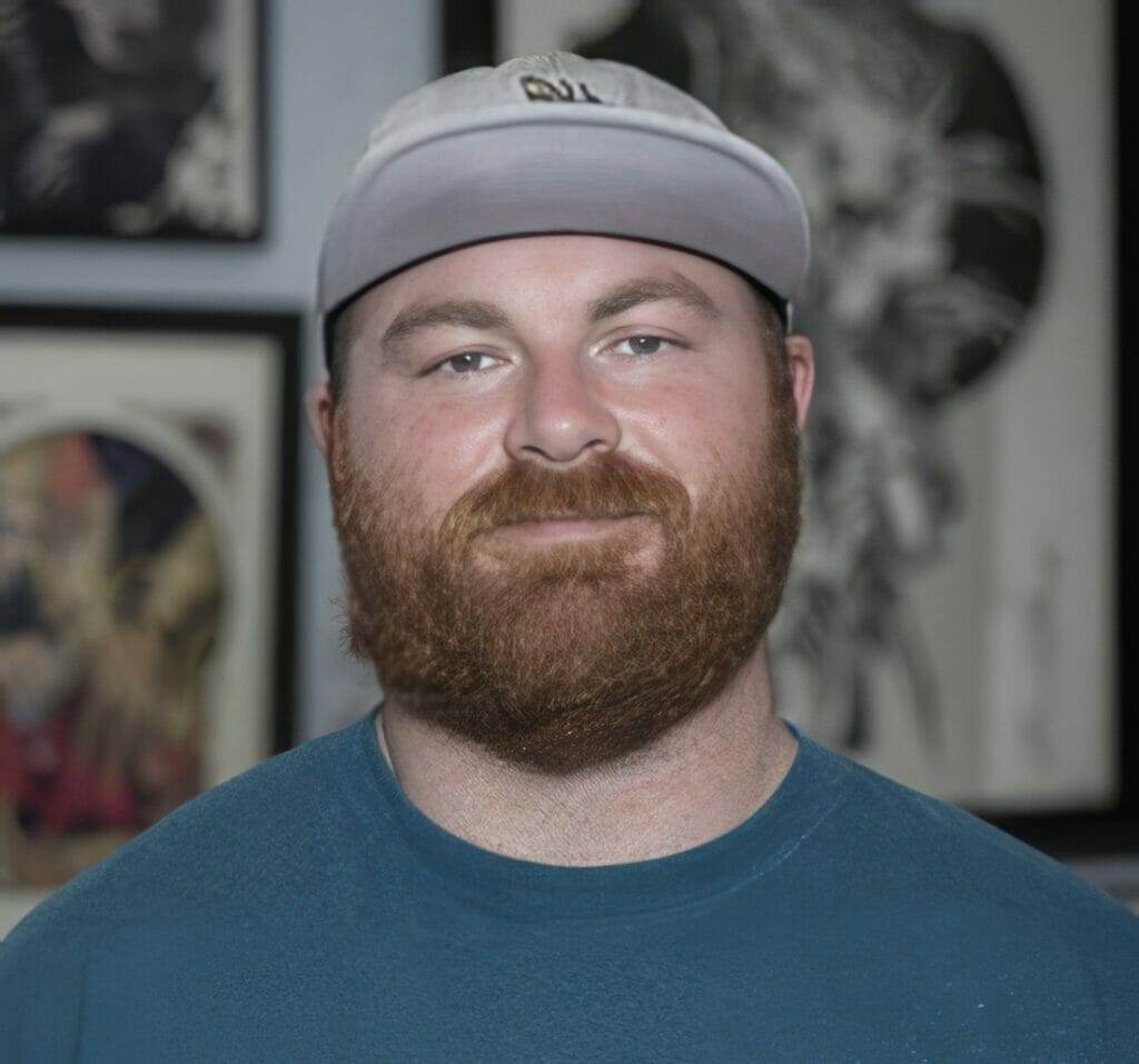 A man with a beard standing in front of framed tattoo pictures at the best tattoo shop in San Diego.