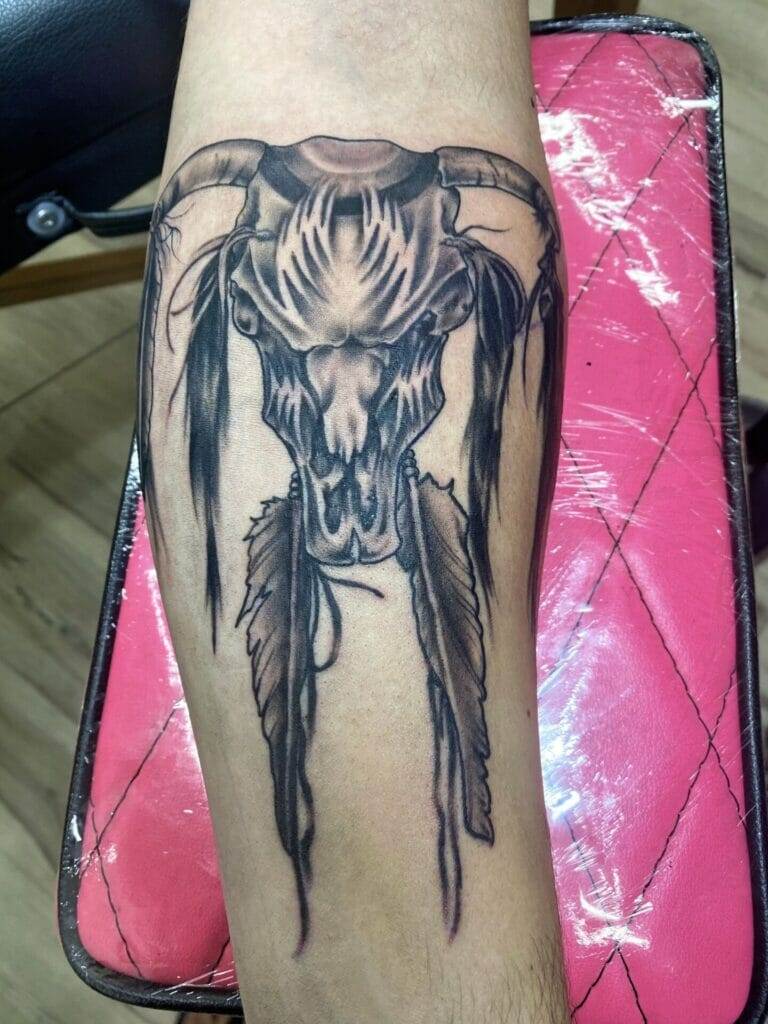 A tattoo of a cow skull with feathers on the forearm.