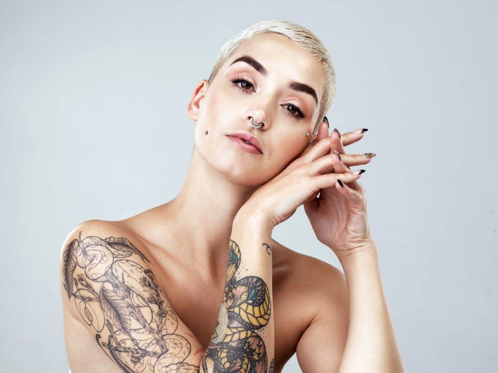         A woman with ink on her arm reviving it with a touch-up, while posing on a gray background.