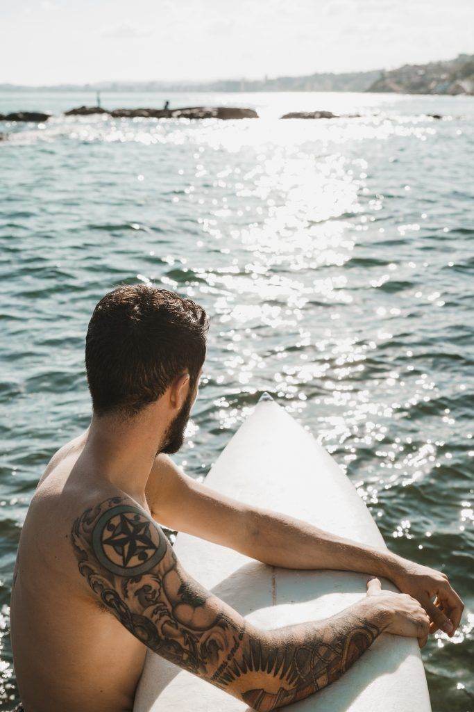 Man with blackwork tattoos sitting on a surfboard at Pacific Beach, looking out over a sunlit ocean.