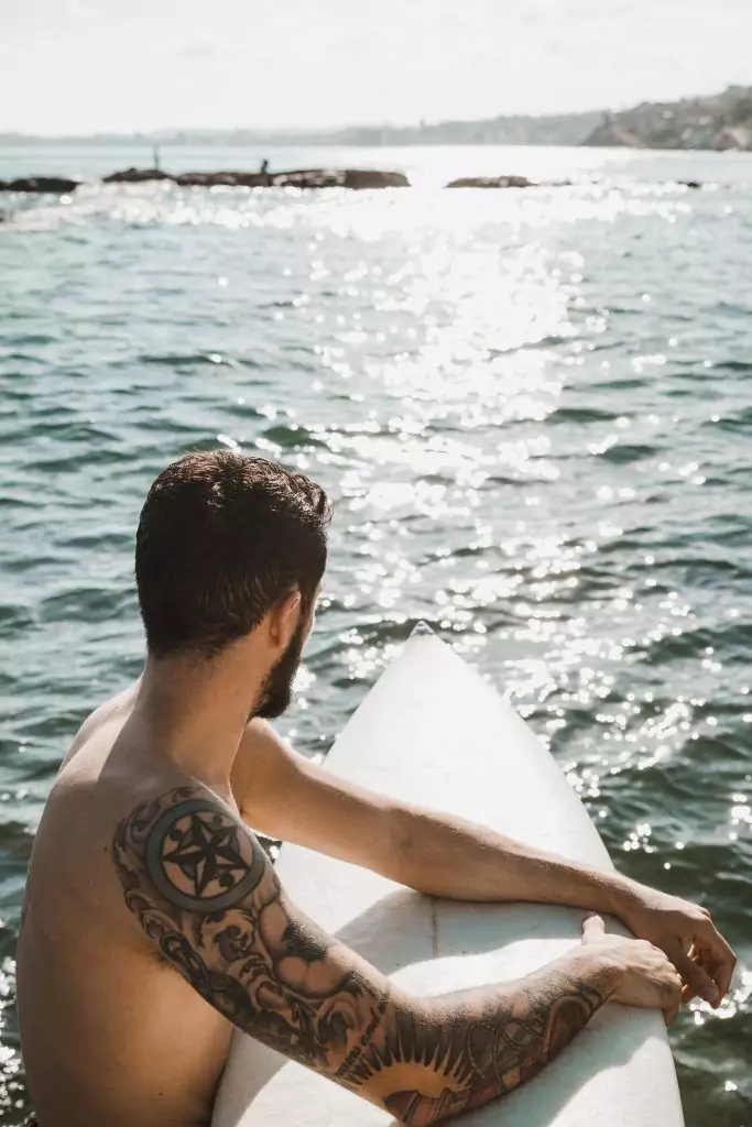 Man with blackwork tattoos sitting on a surfboard at Pacific Beach, looking out over a sunlit ocean.