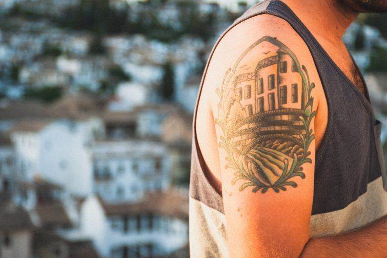 A close-up of a person's upper arm with an illustrative tattoo of a house and landscape framed by leaves. The background is blurred and shows a town with white buildings, evoking the charm of San Diego.