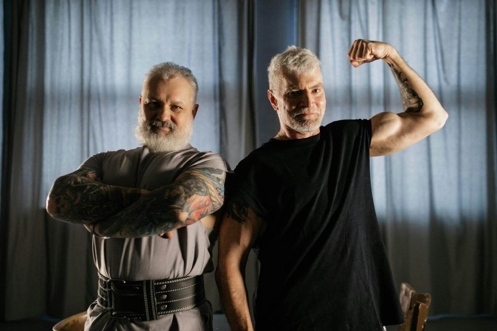 Two older men with white hair and beards, showcasing tattoos and the fascinating ink evolution that comes with age, stand in front of a curtain. One man has arms crossed, while the other flexes his bicep.