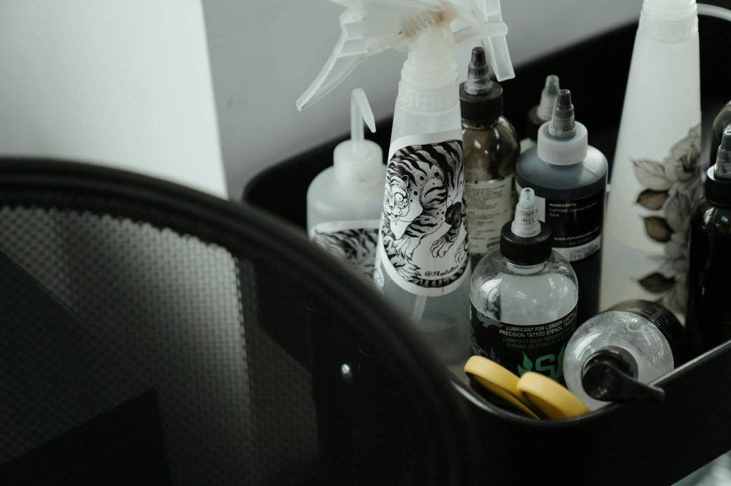 A collection of tattoo ink bottles and a spray bottle with a tiger decal are organized in a black container beside a mesh chair, reflecting the cleanliness essential to professional tattoo studios.