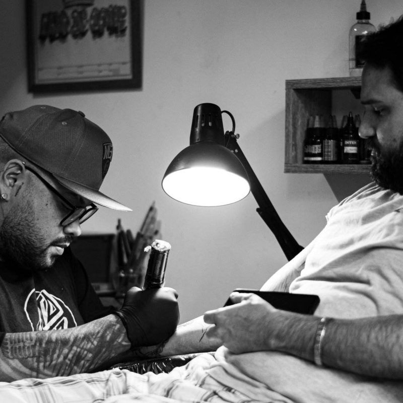 A tattoo artist with a cap and gloves works on a client's arm in a small studio, both focused, under the lamp illuminating the workspace—highlighting the importance of cleanliness in tattoo studios.