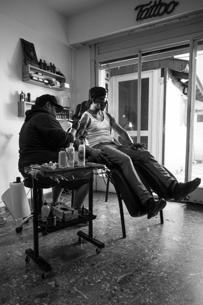 A tattoo artist in a hoodie and cap is tattooing a man in a tank top and hat who is reclining in a chair. Tattooing equipment is visible nearby, ensuring the importance of cleanliness for a safe tattoo environment.