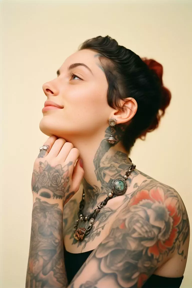 A person with short dark hair and multiple new age tattoos on their arms, neck, and chest, wearing ornate jewelry, looks to the side with a slight smile.