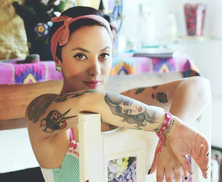 A person with San Diego tattoos on their arms, sporting a headband, sits with relaxed confidence, their arms draped over the back of a chair. In the backdrop, colorful cloth and objects create a vibrant atmosphere.