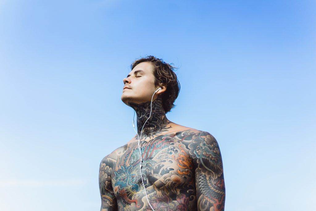 A tattooed man sporting a Dotwork Tattoo, with his eyes closed, enjoys the sunshine against a clear blue sky.