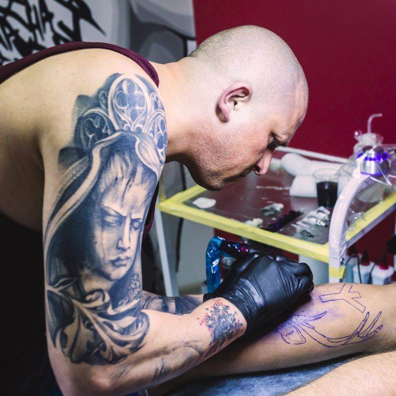 In a San Diego tattoo studio, a tattoo artist with arm tattoos skillfully focuses on creating intricate designs on a person's leg.