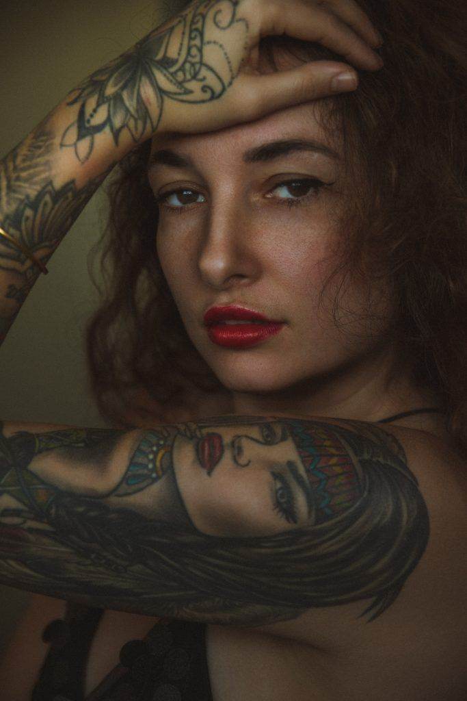 A person with curly hair and bright red lipstick poses with a hand on their forehead at Pacific Beach, showcasing arm tattoos from Funhouse Tattoo, including a large portrait of a woman with a headband.