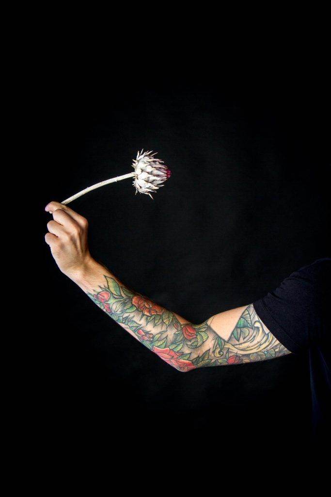 A tattooed arm, adorned with intricate Stick and Poke Tattoos, holds a dried thistle flower against a dark background.