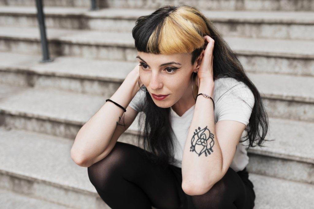 A woman with a black and blonde hairstyle sits on concrete steps in San Diego, wearing a white shirt and black pants. She has a New Age Tattoo on her right arm and is holding her head with both hands.