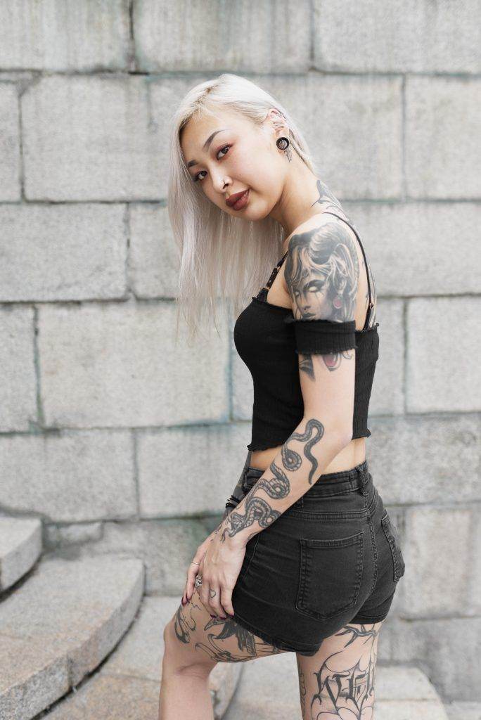 A person with long white hair and visible Funhouse Tattoo designs on their arms and legs stands on stone steps near a gray brick wall, dressed in a black crop top and shorts.