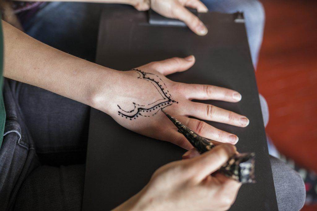 A person is meticulously applying an intricate henna design on the back of another person's hand using a cone applicator, reminiscent of the fine line tattoos found at Funhouse Tattoo.