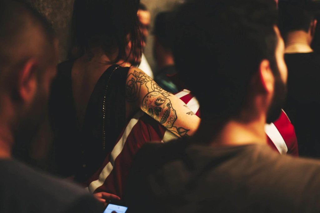 A crowd of people, with a focus on an individual with a tattooed arm. The tattoo features a skull. Immersed in the vibrant culture, the person in the foreground uses a phone.