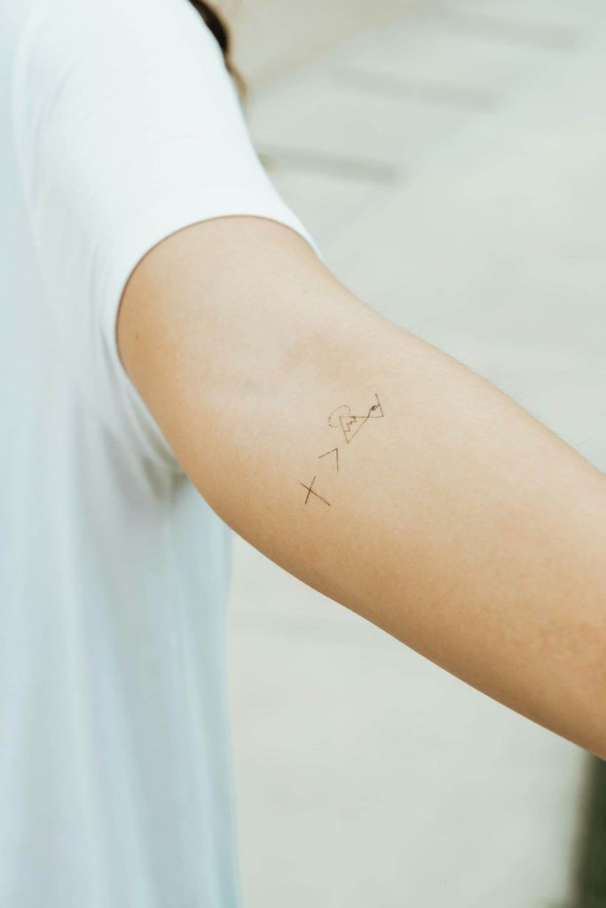 A close-up of a person's arm with a small tattoo featuring symbols, including an 'X', 'Y', and other geometric shapes, showcases the role of unique designs in the tattoo industry. The person is wearing a white short-sleeve shirt, perfect for sharing on social media.