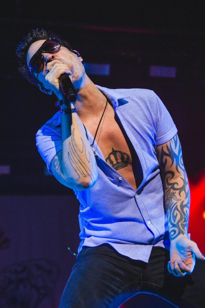 A man with tattoos, wearing a blue shirt and sunglasses, sings passionately into a microphone on stage, embodying the impact of music and tattoo culture against a backdrop of colorful lighting.