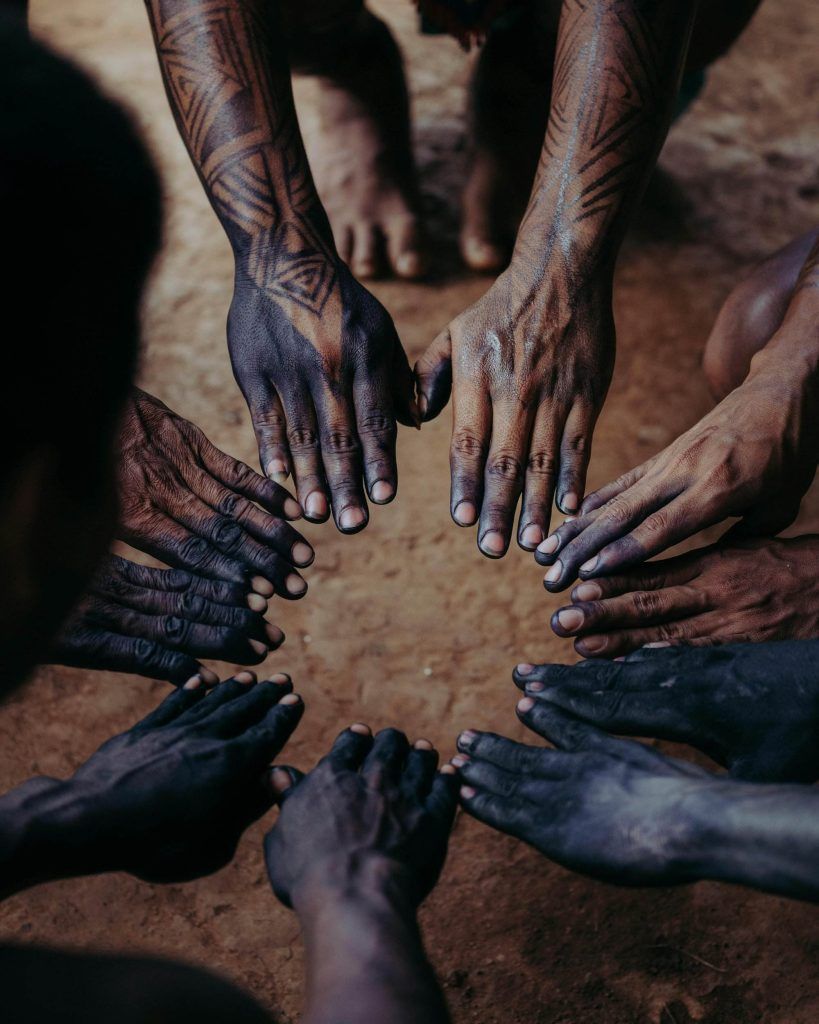 A group of people with tribal tattoos on their forearms and painted hands form a circle on a dirt ground, touching their fingertips together, each design bearing cultural significance and stories behind the ink.