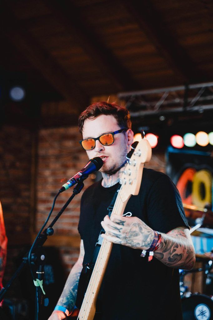 A tattooed musician, embodying the spirit of tattoo culture, wears sunglasses as he plays bass guitar and sings into a microphone on stage during a live performance.