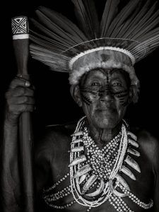 Black and white portrait of an elderly person wearing a feathered headdress, face paint, and multiple beaded necklaces, holding a staff with hand-carved details. Intricate tribal tattoos adorn their arms, each marking stories behind the ink and showcasing deep cultural significance.