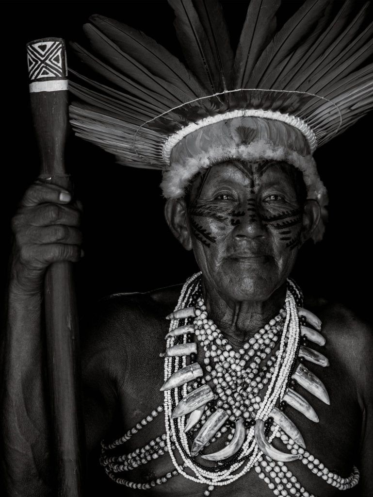 Black and white portrait of an elderly person wearing a feathered headdress, face paint, and multiple beaded necklaces, holding a staff with hand-carved details. Intricate tribal tattoos adorn their arms, each marking stories behind the ink and showcasing deep cultural significance.