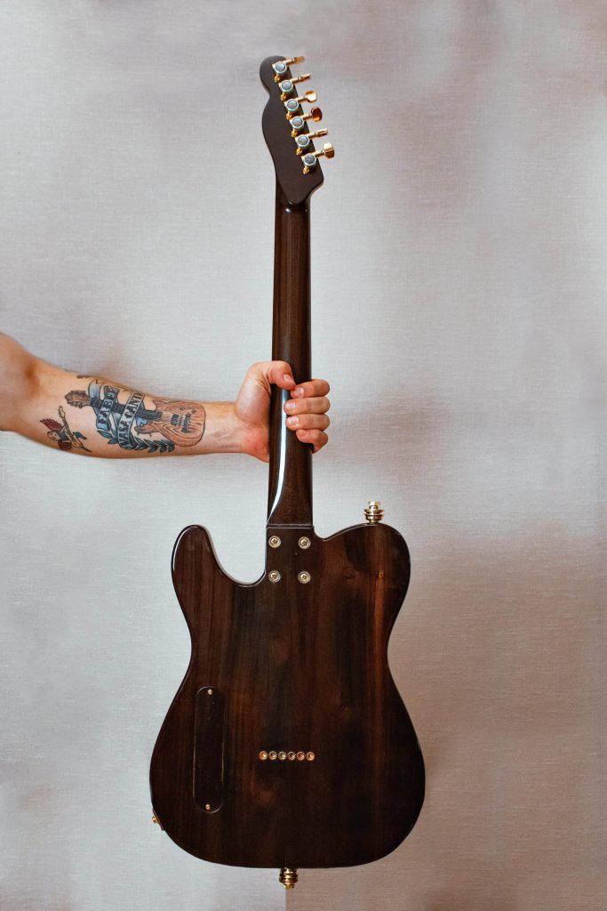 A person with tattoos on their arm holds up a dark wood electric guitar by its neck against a plain background, embodying the powerful impact of tattoo culture in music.