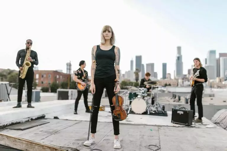 A band performs on a rooftop with a cityscape in the background. A woman holding a violin, her arm adorned with a tattoo, stands in the foreground, with musicians playing saxophone, guitar, and drums behind her. The scene captures the vibrant culture of urban music.